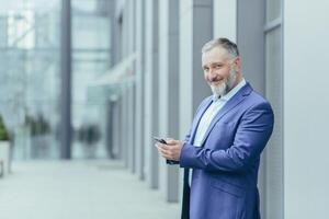 Sénior Beau aux cheveux gris homme des stands dans une costume contre le Contexte de une moderne bâtiment, les usages le téléphone, chèques appels, poster, messages. il regards à le caméra, sourit. photo