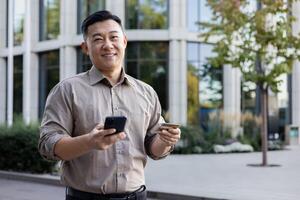 portrait de une Jeune asiatique homme permanent sur le rue, en portant une téléphone et une crédit carte, souriant et à la recherche à le caméra. photo