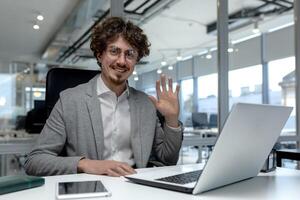 de bonne humeur Jeune homme d'affaire avec frisé cheveux agitant et souriant dans une brillant, moderne Bureau paramètre, engageant dans une virtuel affaires réunion. photo