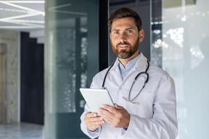 portrait de Sénior mature expérimenté médecin, homme dans médical manteau à l'intérieur clinique à la recherche à caméra, Sénior médecin en portant tablette ordinateur, pour en ligne éloigné consultation de les patients. photo