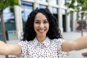 radiant Jeune Latin américain femme d'affaires prise une selfie avec une joyeux expression à l'extérieur une moderne Bureau bâtiment dans le ville. photo