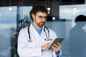 mature réussi médecin à l'intérieur médical bureau, clinique bureau, homme dans blanc médical manteau et stéthoscope les usages tablette ordinateur à consulter les patients à distance en ligne. photo