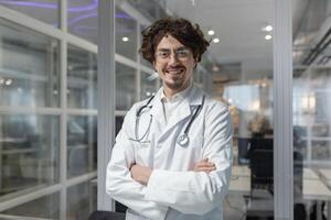 une sur de soi Masculin médecin dans une blanc manteau et stéthoscope permanent avec franchi bras à l'intérieur une médical bureau. homme à la recherche caméra sourire content photo