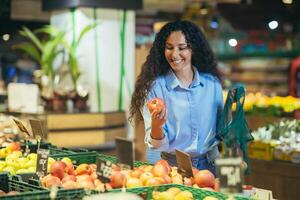 une Jeune hispanique femme est achats dans une supermarché. choix et détient un pomme, met dans un sac écologique photo
