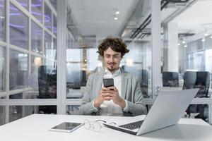 une concentré Masculin Bureau ouvrier avec frisé cheveux en utilisant une téléphone intelligent, portable dans de face de lui, à l'intérieur une contemporain Bureau paramètre. photo