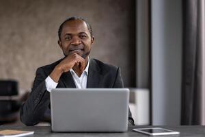 une professionnel noir homme d'affaire sourit en toute confiance dans une moderne Bureau paramètre, en utilisant une portable à le sien bureau entouré par technologie. photo