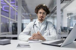 une médecin dans une blanc manteau et stéthoscope séance à une bureau à l'intérieur une médical bureau, en utilisant une portable. homme sérieux à la recherche caméra, portrait photo