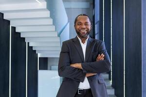 portrait de mature et réussi homme d'affaire à l'intérieur bureau, patron souriant et à la recherche à caméra avec franchi bras, africain américain homme dans affaires costume content avec réussi réussite résultats. photo