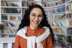 portrait de attrayant Jeune femme avec frisé cheveux portant lunettes et décontractée tenue sur flou librairie Contexte. femelle en train de lire amoureux visite préféré magasin pour fiction Littérature achat. photo