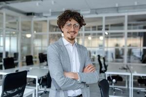 portrait de une Jeune mature Masculin professionnel avec frisé cheveux en toute confiance permanent dans une bien éclairé entreprise bureau. photo