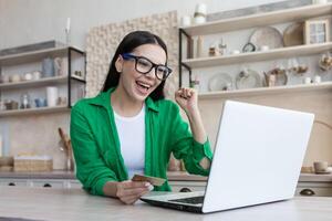 Jeune magnifique femme dans des lunettes et une vert chemise est occupé travail sur une portable à Accueil cuisine photo