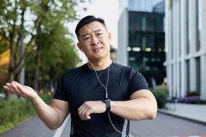 portrait de dérangé Jeune asiatique homme fonctionnement dans écouteurs sur le des rues de le ville, permanent à la recherche sérieusement à le caméra, diffusion le sien mains, mécontent avec le résultat sur le intelligent montre. photo