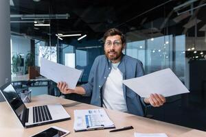 mécontent et en colère homme d'affaire à l'intérieur Bureau montrant rapports et factures les documents à caméra, patron dans chemise à la recherche à caméra travail avec portable papier travail. photo