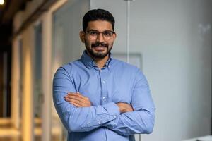 homme d'affaire avec franchi bras souriant et à la recherche à caméra, portrait de content Masculin ouvrier à l'intérieur bureau, patron dans des lunettes et chemise, content avec réussite résultats, fier et confiant. photo