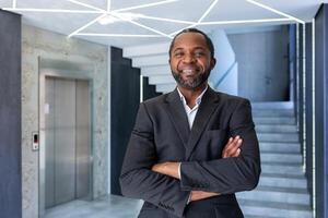 portrait de mature et réussi homme d'affaire à l'intérieur bureau, patron souriant et à la recherche à caméra avec franchi bras, africain américain homme dans affaires costume content avec réussi réussite résultats. photo