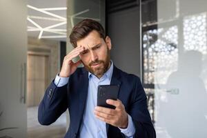 fermer photo de une sous le choc homme permanent dans une costume dans le Bureau et en train de lire mal nouvelles et messages, en portant le sien tête dans choc.