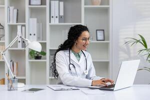 souriant latina médecin dans sa Bureau avec stéthoscope, travail Heureusement sur portable dans une santé se soucier paramètre. photo