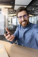 content barbu homme dans une bleu chemise atteindre vers le caméra, offre une salutation avec une chaud, amical sourire dans un Bureau paramètre. photo