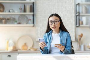 portrait de une Jeune magnifique asiatique femme en portant une crédit carte et une téléphone dans sa main. séance à Accueil dans le cuisine, à la recherche à le caméra. photo
