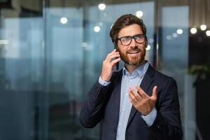 mature homme d'affaire dans une affaires costume parlant sur le téléphone, le directeur est satisfait avec le résultat de le réussite et travail pourparlers à propos le positif résultat, le homme est à travail à l'intérieur le bureau. photo