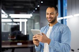 une professionnel homme d'affaire dans décontractée tenue est sur la photo souriant tandis que se concentrer sur une numérique tablette dans une contemporain Bureau environnement. le atmosphère transmet connectivité, productivité, et La technologie à le lieu de travail.