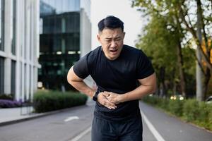 asiatique Jeune Masculin athlète courbé plus de de douleur dans le sien côté, en portant le sien estomac avec le sien mains après fonctionnement autour le ville. photo