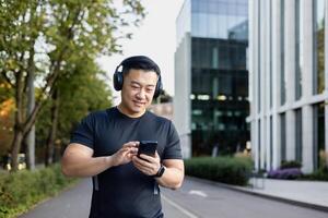 une Jeune asiatique homme dans tenue de sport est permanent sur une ville rue portant écouteurs et en utilisant une mobile téléphone. photo