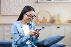 une content Jeune magnifique asiatique femme est séance sur le canapé à maison. elle est en portant une mobile téléphone dans sa mains, l'écriture une message. correspond avec petit ami, amis, sourit photo