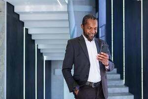 africain américain souriant homme d'affaire, exécutif directeur, avocat permanent dans hall de Bureau centre dans affaires costume, en portant main dans poche et en utilisant téléphone. photo