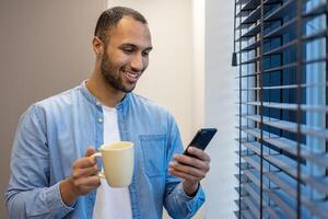 un Indien homme dans une décontractée denim chemise jouit le sien Matin café tandis que en utilisant le sien téléphone intelligent dans le vivant chambre. photo