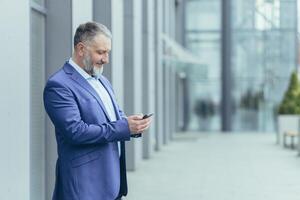 Sénior Beau homme d'affaire homme des stands sur le rue près le Bureau centre et attend pour clients, employés, détient le téléphone dans le sien mains, cadrans une nombre, messages. photo