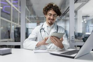 une médecin portant une blanc laboratoire manteau et stéthoscope est vu en utilisant une tablette à l'intérieur une médical Bureau à le clinique. homme content sourire en train de lire rapports médical photo