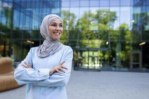 portrait de une positif, réussi musulman femme d'affaires dans une hijab, en toute confiance souriant dans un Urbain paramètre. photo