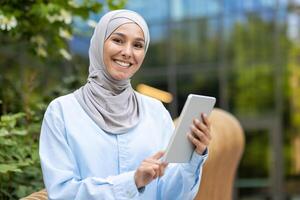 une portrait de une content musulman femme dans une hijab en portant une tablette, exprimer confiance tandis que permanent en plein air avec feuillage et moderne architecture dans le Contexte. photo