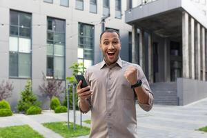 content et sous le choc par Matin nouvelles, arfo-américain homme d'affaire souriant et à la recherche à caméra de à l'extérieur Bureau bâtiment, homme en marchant vers le bas rue en utilisant mobile téléphone photo