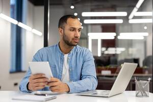 une sérieux Jeune homme, une Danseur, une designer travaux dans le Bureau sur une ordinateur portable, est assis à une table et les usages une tablette dans le sien mains. photo