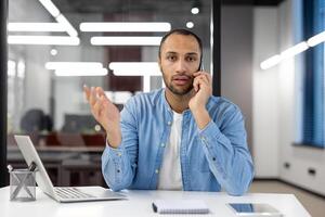 une homme est parlant sur le sien cellule téléphone tandis que séance à une bureau. il est portant une bleu chemise et blanc chemise photo