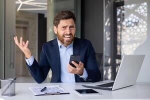 portrait de un en colère Jeune homme d'affaire séance à une bureau dans le bureau, en portant une téléphone dans le sien main et à la recherche déçu à le caméra. photo