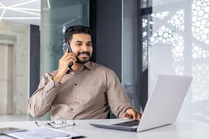 une professionnel Jeune homme d'affaire bavardage sur le téléphone tandis que travail sur le sien portable dans une moderne Bureau environnement, convoyer une sens de affaires la communication et productivité. photo