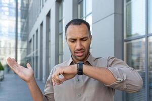 une homme est à la recherche insatisfait à une montre intelligente. une Latin américain homme d'affaire est attendre à l'extérieur un Bureau bâtiment photo