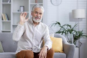 de bonne humeur personnes âgées homme dans une décontractée chemise fabrication une appel, agitant à le caméra avec une chaud sourire dans une confortable Accueil paramètre. photo