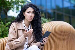 dérangé hispanique femme avec frisé cheveux séance sur une banc à l'extérieur en portant une téléphone dans sa main et à la recherche à le caméra déçu, Jeune brunette eu mal nouvelles, problèmes à travail. photo