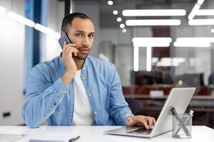 une concentré homme dans décontractée affaires tenue engagé dans une téléphone conversation tandis que simultanément travail sur le sien portable dans une moderne Bureau environnement. photo