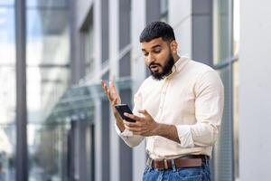 en colère Jeune Indien homme permanent sur le rue près une bâtiment et à la recherche frustré à le mobile téléphone filtrer, faire des gestes avec le sien mains émotionnellement photo