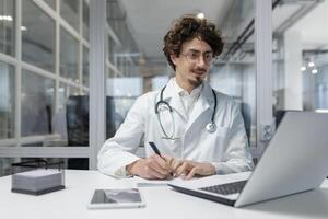 une dévoué Masculin médecin dans une blanc manteau et stéthoscope travail à le sien bureau en utilisant une portable. homme sourire content l'écriture rapport photo