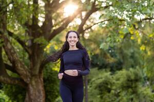 Jeune magnifique femme fonctionnement dans le parc, portrait de une Latin américain femme le jogging et Faire actif des exercices, sportive souriant et à la recherche à le caméra parmi le des arbres. photo