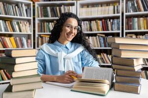 une de bonne humeur Jeune femme avec des lunettes et frisé cheveux études parmi piles de livres dans une bibliothèque, exsudant curiosité et enthousiasme. photo