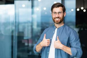 portrait de homme d'affaire dans décontractée chemise, homme avec barbe et des lunettes souriant et à la recherche à caméra montrant les pouces en haut affirmative, affaires propriétaire travail à l'intérieur bureau. photo