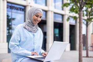 une de bonne humeur musulman femme dans une bleu hijab travaux sur sa portable tandis que séance à l'extérieur une contemporain bâtiment. elle est enthousiaste et concentré, mettant en valeur la diversité et la modernité dans travail environnements. photo