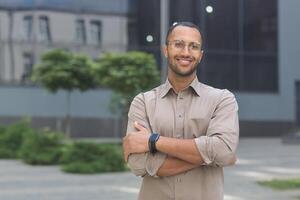 portrait de Jeune hispanique étudiant homme souriant et à la recherche à caméra près Université Campus avec bras franchi portant des lunettes et chemise photo